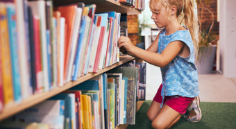 Una niña pequeña examina libros en una gran estantería, en el entorno de una biblioteca.