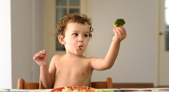 Un niño pequeño está sentado a la mesa sin camiseta, sosteniendo un trozo de brócoli en el aire, con un plato de comida sobre la mesa.