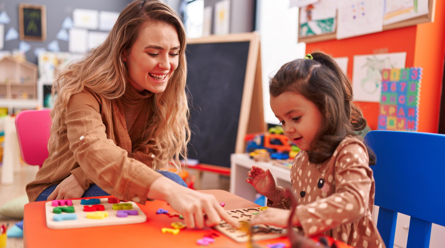 Una mujer adulta sentada con un niño pequeño en un aula colorida, ayudando al niño con un rompecabezas.
