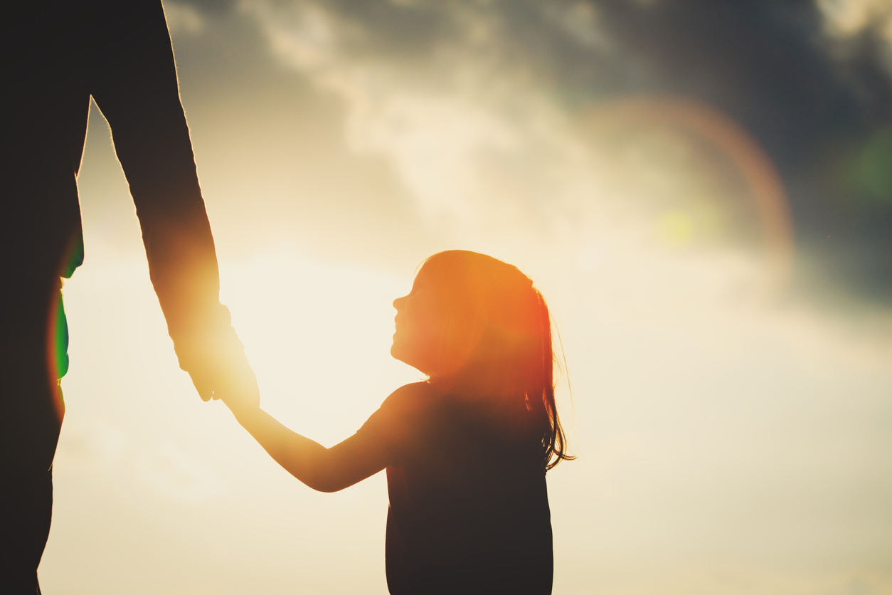 Foto a contraluz con la silueta de una niña pequeña tomada de la mano de una persona adulta.