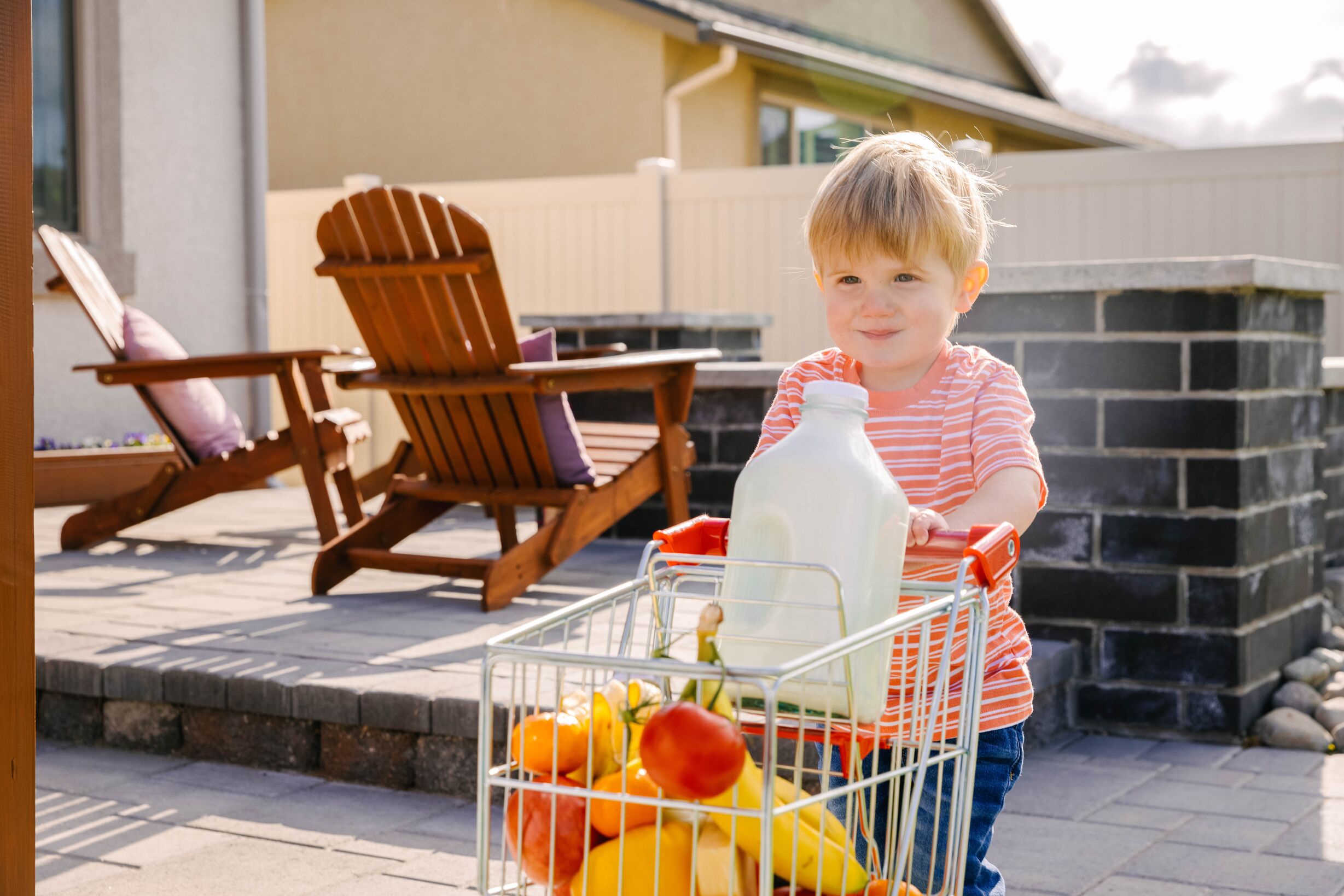 Un niño pequeño que viste una camisa de rayas naranjas empuja un mini carrito de compras con frutas y un gran recipiente de leche.