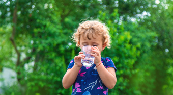 Un niño pequeño bebe agua de un pequeño vaso de vidrio mientras está afuera con un fondo de árbol verde brillante.